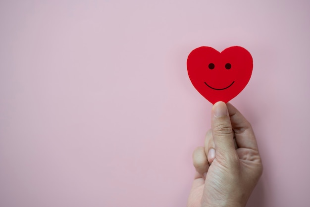 Hands holding red heart with smiley face icon on pastel pink\
background