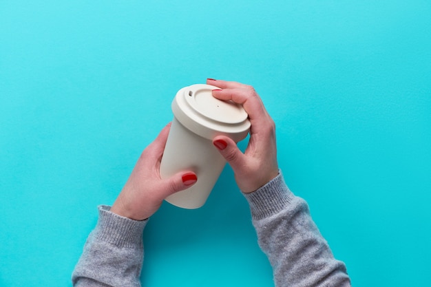 Photo hands holding reusable coffee cup made from eco friendly bamboo with silicone lid. eco friendly zero waste tea mug on blue mint paper without plastic. travel solution for sustainable lifestyle.