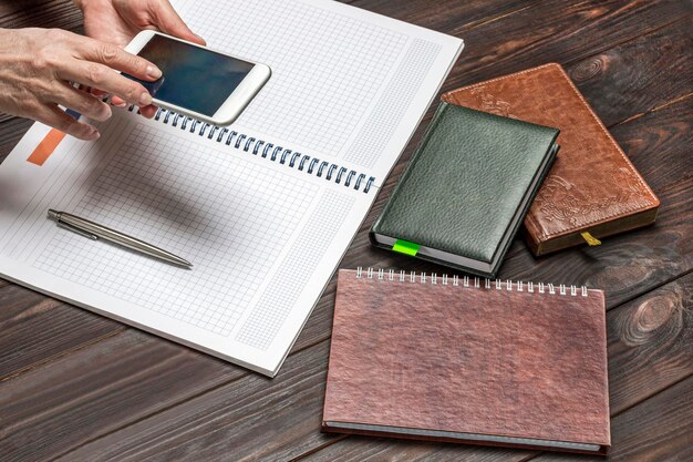 Hands holding selfphone over an open notebook Calculator and notebooks on table Work from home Dark wooden background Top view