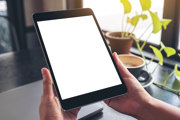  hands holding and using black tablet pc with blank white desktop screen with notebook, green leaves and coffee cup on wooden table