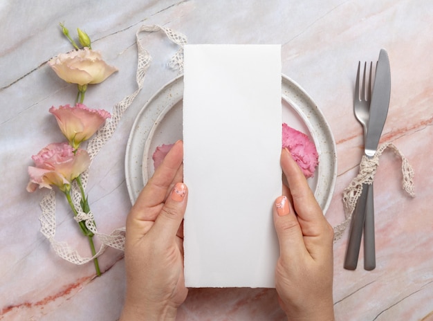 Руки держат свадебное меню над керамической тарелкой на мраморном столе, украшенном цветами и лентами. сцена макета с карточкой меню чистого листа бумаги. женственная элегантная плоская планировка