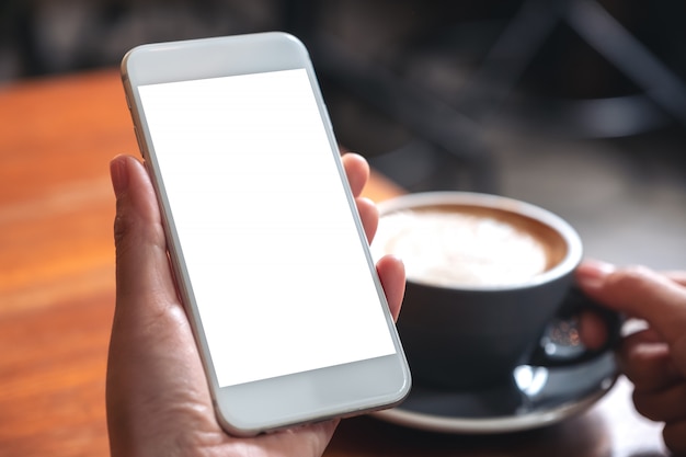 hands holding white mobile phone with blank screen while drinking coffee