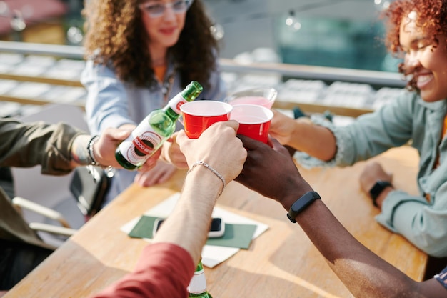 Hands of interracial friends clinking with drinks ove table in outdoor cafe