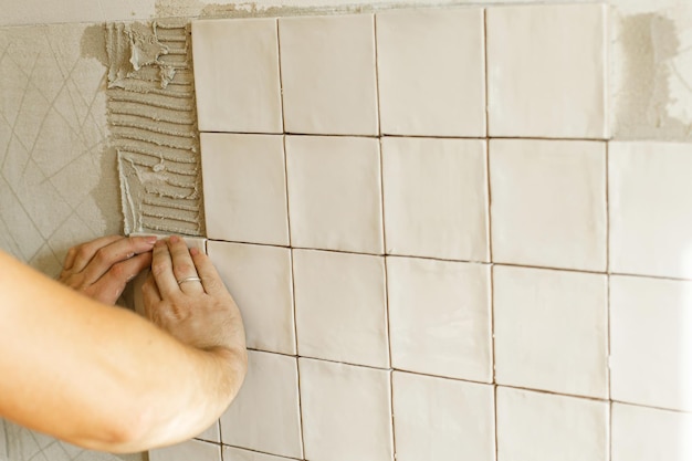 Hands laying modern square tile on adhesive close up Worker installing stylish white tiles on plaster wall Renovation