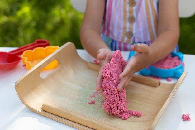 Hands of little girl playing with colorful kinetic sand close-up. Leisure activity outdoors in summer day. Crafting, modeling, creating. Creative hobby. Pink color