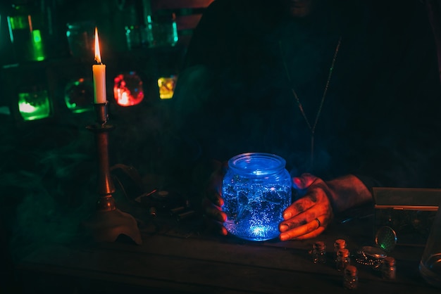 Hands of magician alchemist scientist with a flask with a magic potion at table in mystical laboratory