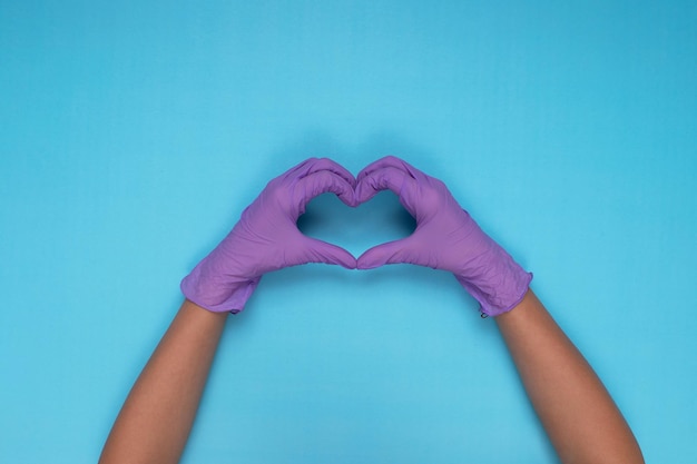 Hands making heart sign with lilac colored medical gloves on light blue background