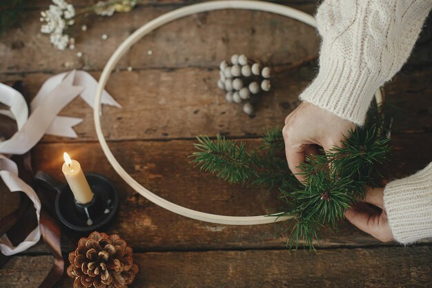 Hands making modern christmas wreath on rustic table with fir
branches wooden hoop candle