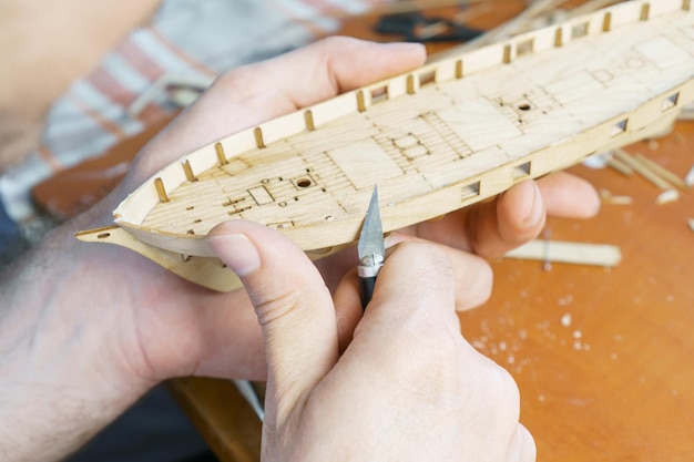 Hands man cutting out details for ship model with clerical\
knife from plywood process of building toy ship handicraft