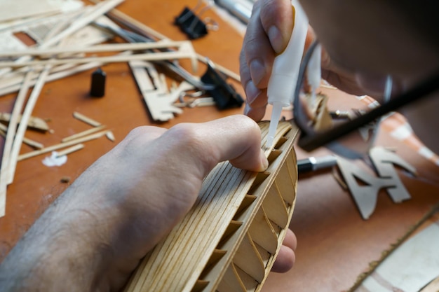 Hands of man gluing plywood details for ship model with glue holding with fingers Process of building toy ship