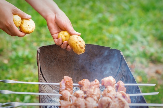 人間の手は、屋外で火をつけてグリルで串に刺したジャガイモを使ったバーベキュー肉を準備します。ライフスタイルの素朴な食事の準備の概念