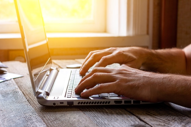 Hands of a man working at a computer laptop by the window in the sunlight