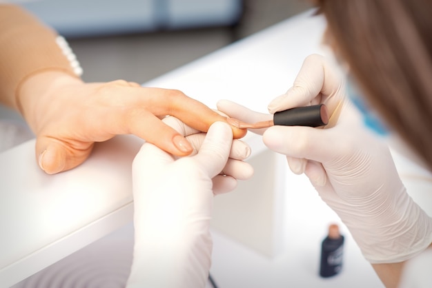 Le mani del maestro di manicure in guanti protettivi bianchi applicano lo smalto alle unghie femminili.