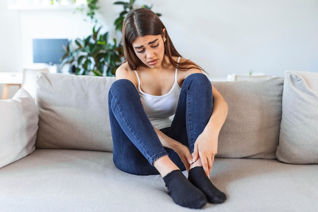 Photo hands massaging swollen foot while sitting on sofa during the day at home. photo of young caucasian woman suffering from pain in leg. woman massaging her legs after all day at work in office
