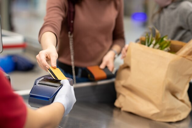 Hands of mature female buyer with smartphone over payment\
machine going to pay for food products in supermarket by cash\
register