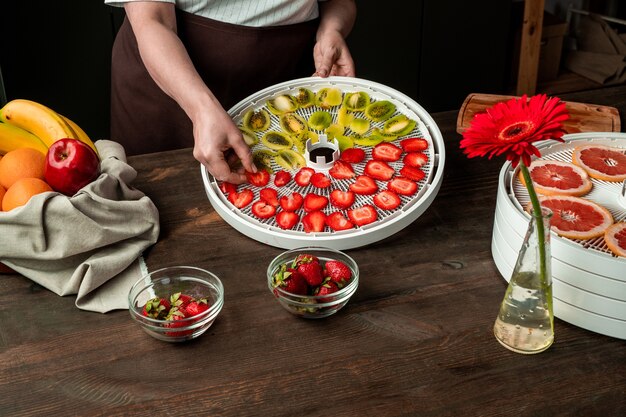 Hands of middle aged woman putting sliced strawberries and kiwi on tray of fruit dryer among fresh bananas, oranges, etc on kitchen table