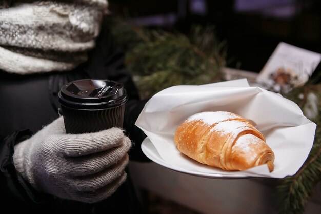 Hands in mittens hold a hot cup of coffee and a croissant. Coffee to go in winter.