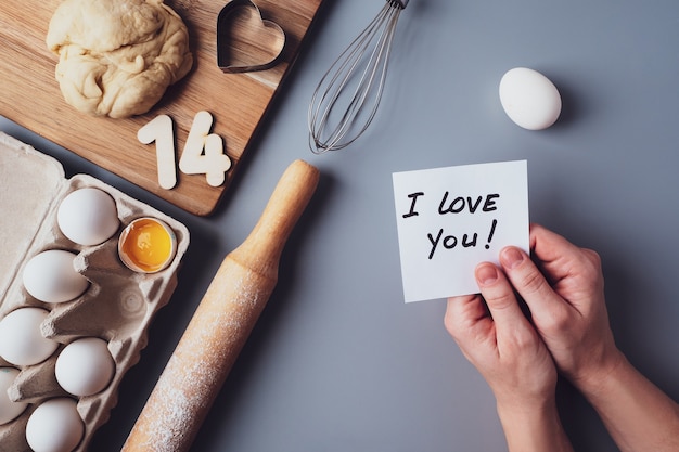 Foto nelle mani di una nota ti amo. ingredienti per fare i biscotti fatti in casa su uno sfondo grigio. il concetto di cucinare dolci per san valentino, festa del papà o festa della mamma. disposizione piana, vista dall'alto.