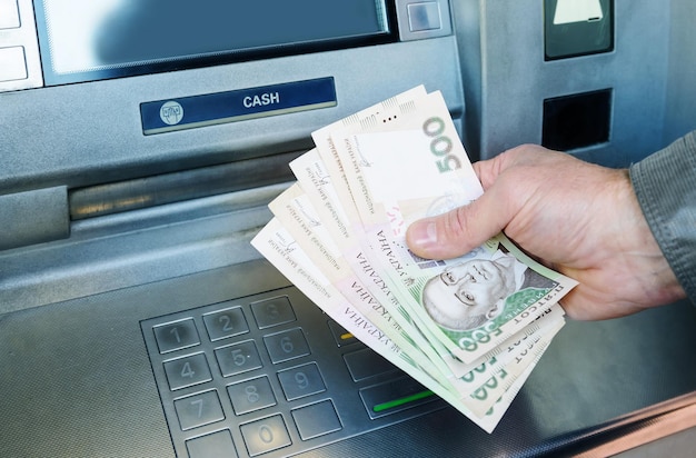 Фото Руки мужчины держат банкноты украинской гривны на фоне банкомата.