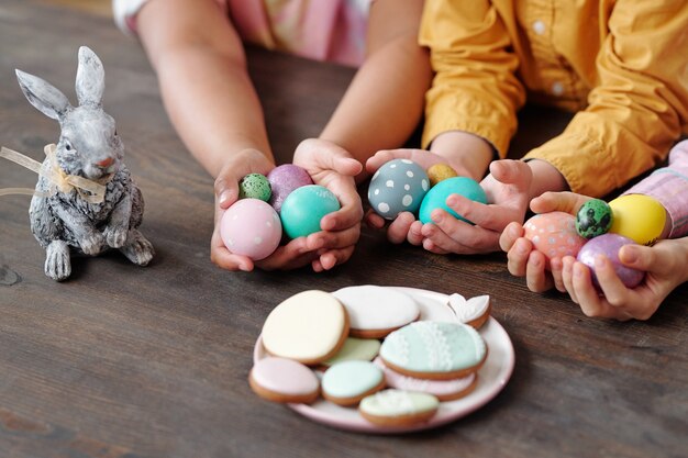 Руки трех межкультурных маленьких детей в повседневной одежде с крашеными пасхальными яйцами среди печенья и игрушечного кролика, сидя за столом