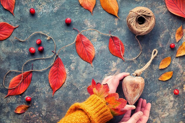 Le mani in maglione arancione tengono il cuore di legno. decorazioni naturali autunnali, rosso vivo, foglie di quercia gialle. lay piatto su sfondo scuro.
