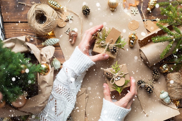 Hands pack a gift for Christmas and new year in eco-friendly materials: kraft paper, live fir branches, cones, twine, tied with a bow. Tags with mock up, natural decor, hand made, flatly. Festive mood