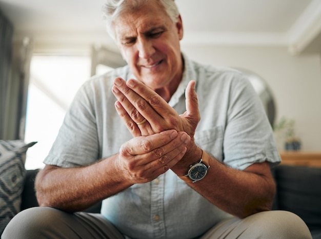 Dolore alle mani e artrite con un uomo anziano che gli tiene la mano mentre soffre di crampi o lesioni da osteoporosi salute medica e articolare con un anziano pensionato maschio nel soggiorno di casa