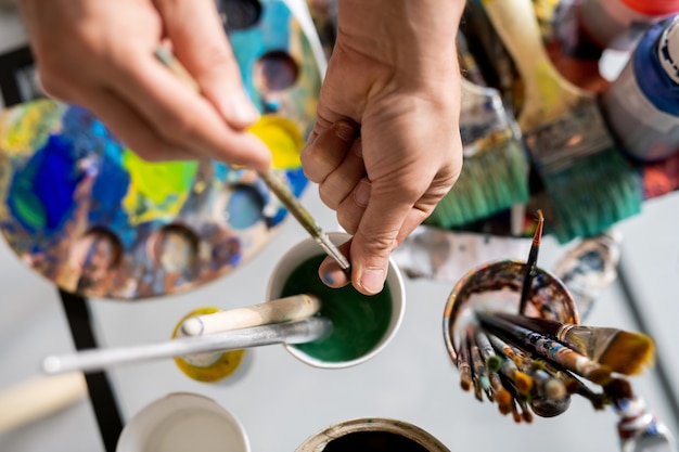 Photo hands of painter holding paintbrush over glass of water while cleaning and drying bristles before work