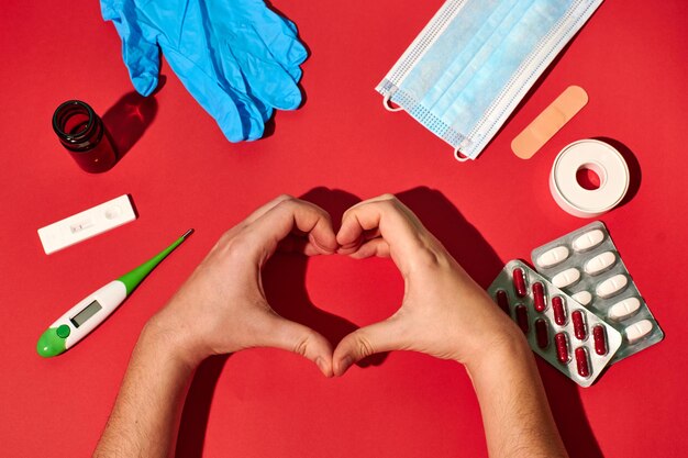 Foto mani di una persona che fa un cuore circondato da oggetti medici su sfondo rosso