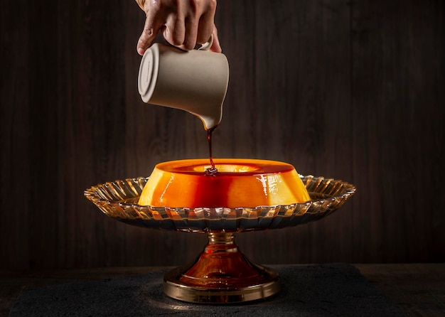 hands pouring sweet syrup over caramel custard pudding on a dark wooden background