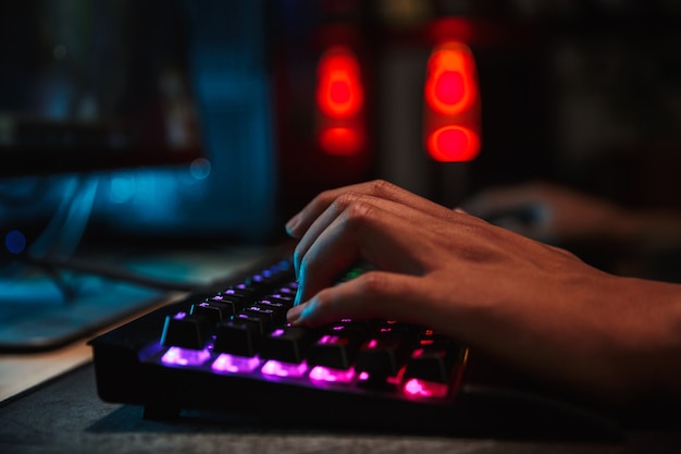 Hands of professional gamer boy playing video games on computer in dark room, using backlit colorful keyboard