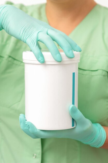 Hands in protective gloves of beautician open a white body cream jar on white background.