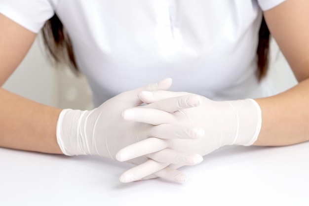 Hands in protective white medical gloves of the woman sitting at the white table. Crossed fingers in gloves