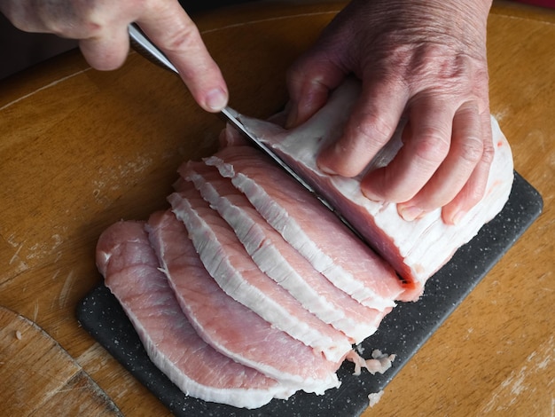 Hands of a seigners woman cut with a knife a raw pork meat fillet on a plastic black board steaks