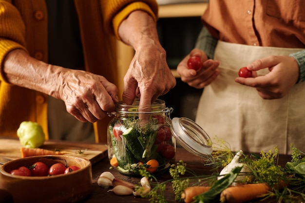 Photo hands of senior female putting garden cucumbers into jar with marinade mix