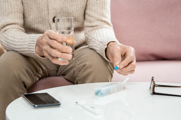 Mani di un uomo anziano malato in abbigliamento casual che tiene pillola e bicchiere d'acqua sul tavolo mentre è seduto sul divano e sta per prendere la medicina