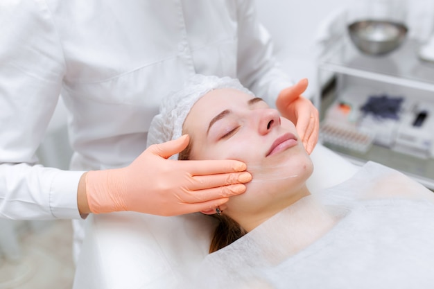 Hands of skillful beautician cleaning and touching female face with cotton pad or sponge in the white office.