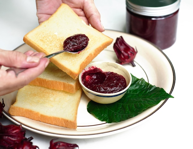 Hands spreading hibiscus jam on a toast against jar with jam and hibiscus flowers