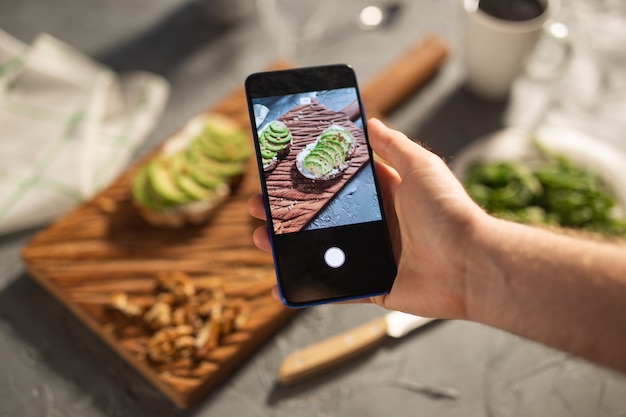 Photo hands take pictures on smartphone of two beautiful healthy sour cream and avocado sandwiches lying on board on the table. social media and food concept