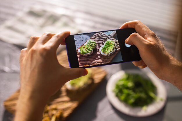 Hands take pictures on smartphone of two beautiful healthy sour cream and avocado sandwiches lying on board on the table. Social media and food concept