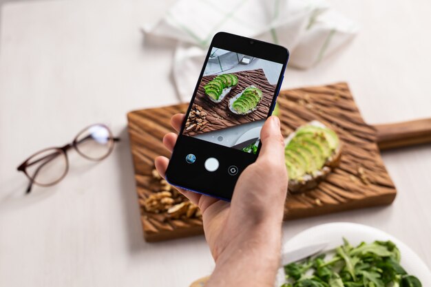 Photo hands take pictures on smartphone of two beautiful healthy sour cream and avocado sandwiches lying