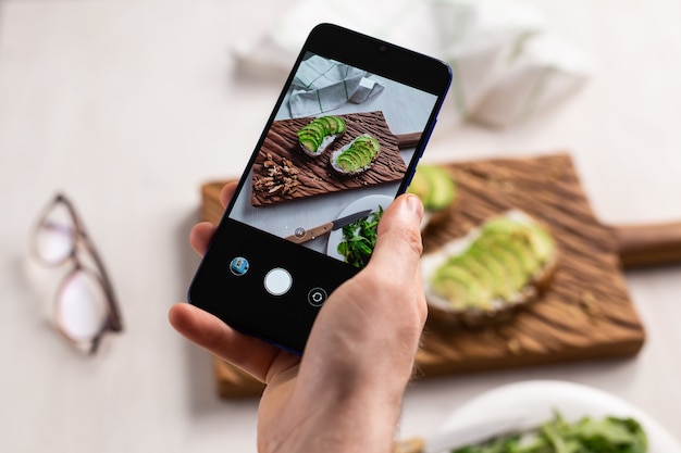 Hands take pictures on smartphone of two beautiful healthy sour cream and avocado sandwiches lying