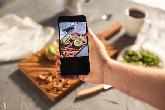 Photo hands take pictures on smartphone of two beautiful healthy sour cream and avocado sandwiches lying