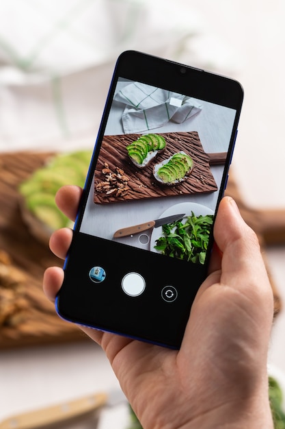 手は2つの美しい健康的なサワークリームとアボカドサンドイッチのスマートフォンで写真を撮ります