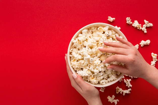 Hands taking popcorn from classic striped bucket on red background. Hot corn scattered from paper box, copy space. Fast food and movie snack, top view