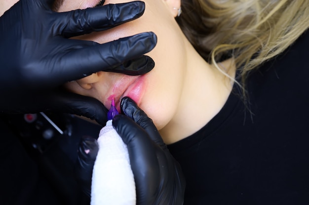Руки татуировщика в черных перчатках держат губы модели и выполняют перманентный макияж губ с помощью тату-машинки.