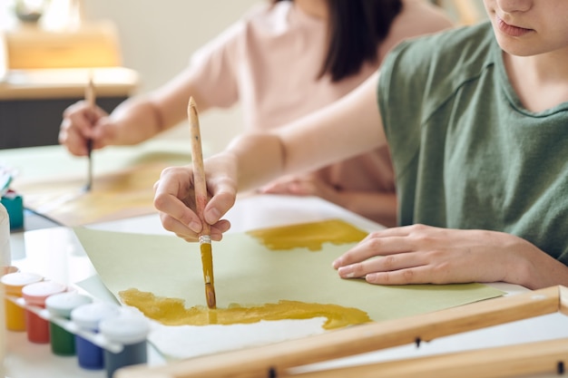 Hands of teenage schoolgirl painting with yellow gouache or watercolor on paper at lesson of moder arts