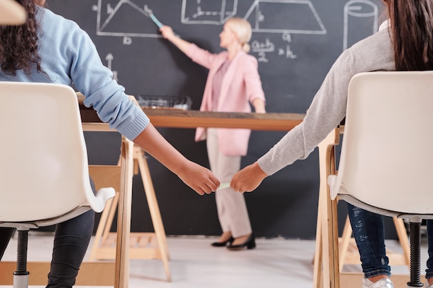 Foto le mani di due studentesse contemporanee sedute ai banchi e che si scambiano culle o appunti stupidi