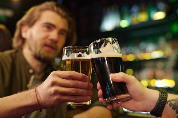 Hands of two friendly guys clinking with glasses of beer in\
front of camera while one of them pronouncing toast against bar\
with alcoholic drinks