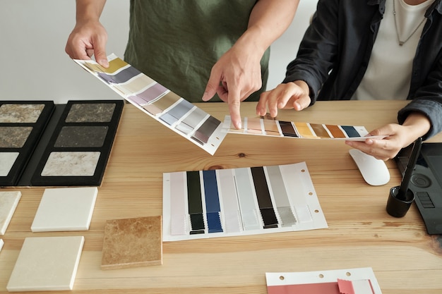 Hands of two young creative designers discussing textile samples while pointing at one of them by workplace